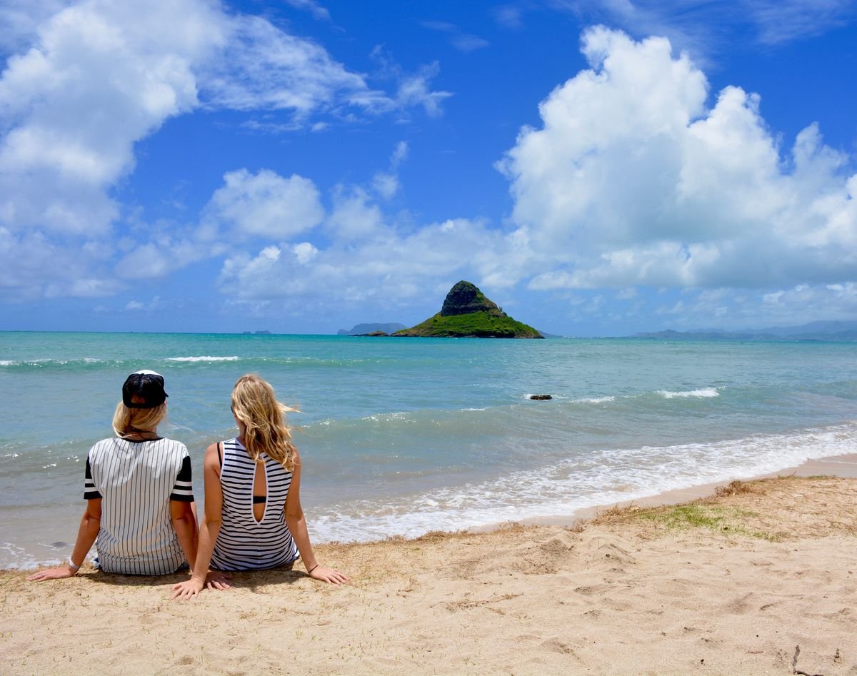 Two visitors on a beach looking out at Mokoli'i island on O'ahu's windward coast